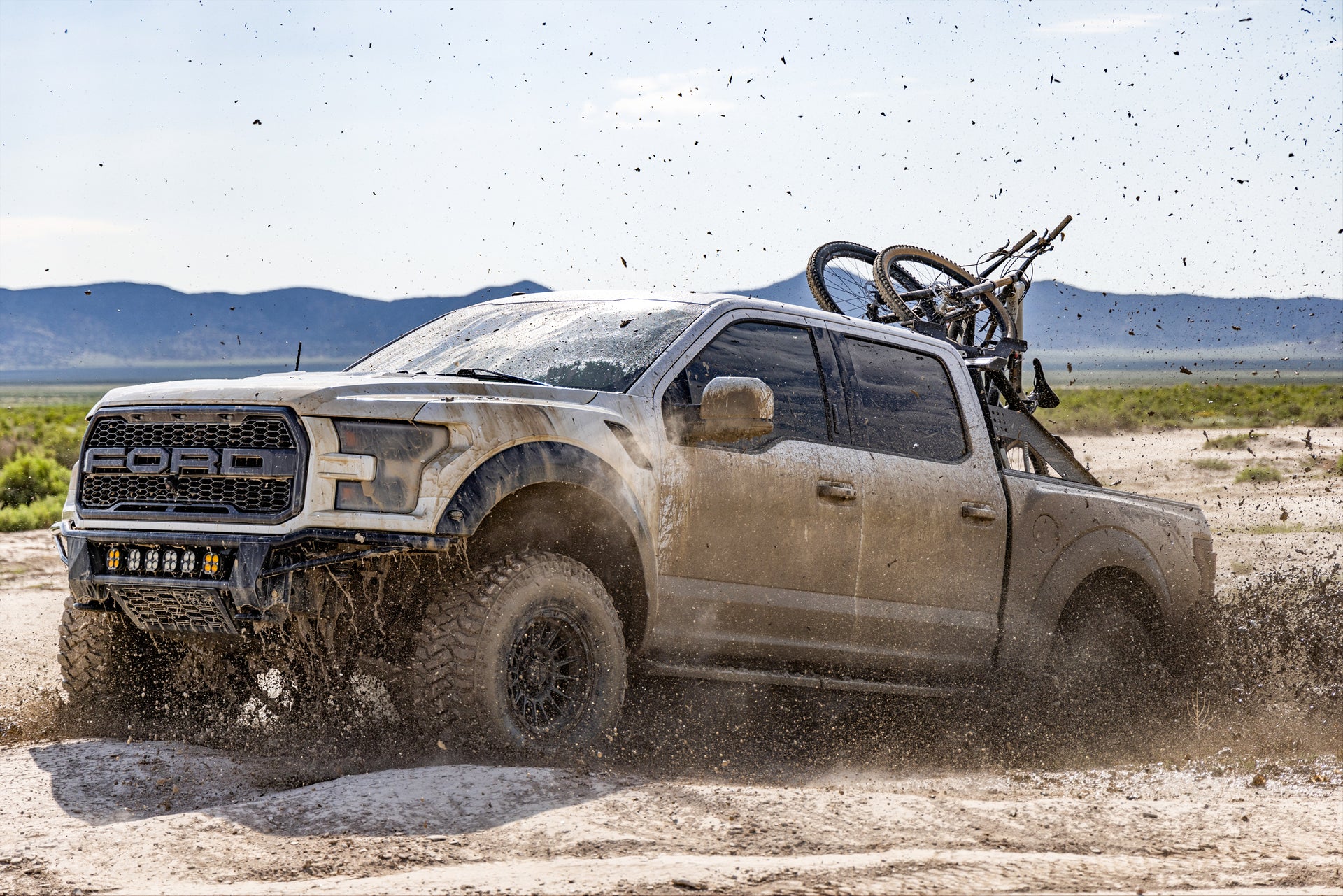 A truck with bikes in the bed going through a huge mud pit doing off roading with mountain bikes in the bed